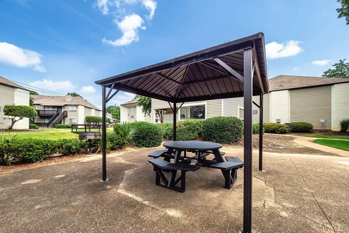Inviting outdoor gazebo with round picnic table set in a serene apartment complex surrounded by lush greenery and clear blue skies.