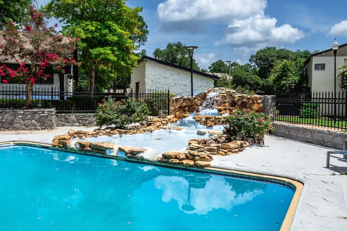 Charming apartment complex pool area with a cascading stone waterfall, vibrant landscaping, and clear blue skies.
