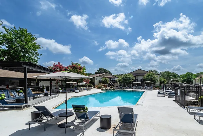 Luxurious apartment complex pool area with modern lounge chairs, umbrellas, and lush greenery under a bright blue sky.