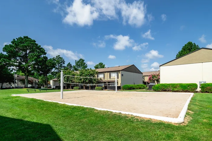 Charming community view featuring a scenic sand volleyball court surrounded by lush greenery and modern apartments under a clear blue sky.