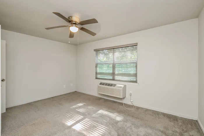Bright and airy bedroom with soft carpeting, large window with blinds, and a ceiling fan, offering serene comfort.
