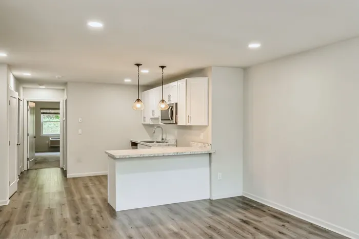 Modern apartment kitchen with sleek white cabinetry, elegant pendant lighting, and a marble countertop, leading to a bright living area with warm hardwood flooring.