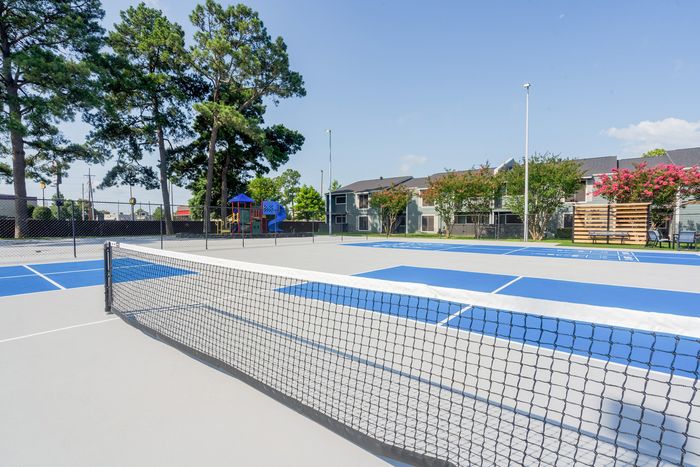 Inviting apartment complex with vibrant blue tennis courts and lush playground amidst serene greenery.
