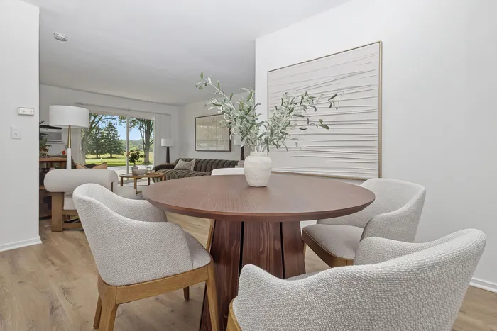 Elegant dining area with a round wooden table and plush chairs, overlooking a bright living space with lush views through large glass doors.