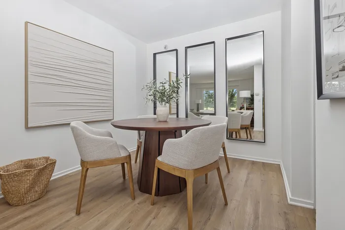 Elegant dining area with a round wooden table, plush chairs, and modern decor, framed by artistic wall accents and full-length mirrors.
