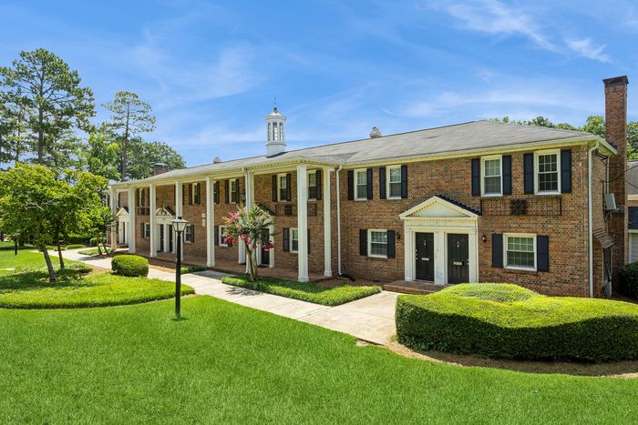 Charming brick apartment building with classic columns, set amidst lush landscaped gardens under a bright blue sky.