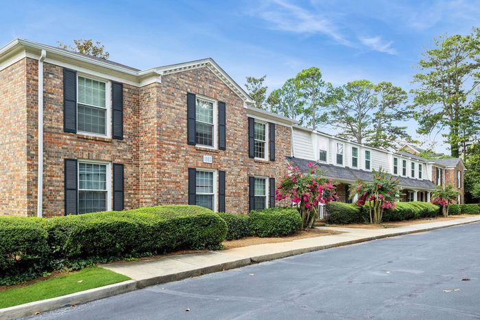Charming brick apartment building with lush green landscaping and vibrant flowering shrubs under a bright blue sky.