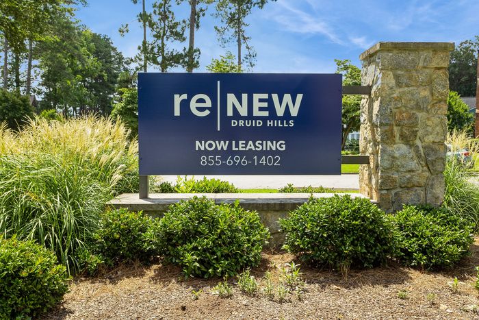 Inviting leasing sign at ReNew Druid Hills, surrounded by lush greenery and clear blue skies, highlighting luxury living.