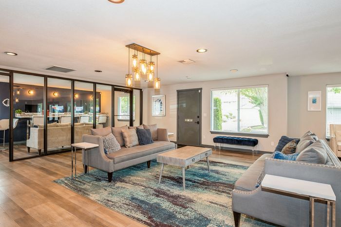 Chic living room with modern decor, featuring a stylish gray sofa set, elegant pendant lighting, and a vibrant area rug. The glass partition gives a glimpse into a sleek office space, creating an open and inviting atmosphere.