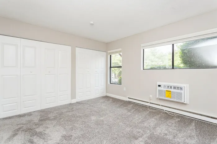 Bright and airy carpeted room with large windows, ample closets, and an air conditioning unit, perfect for comfort and style.