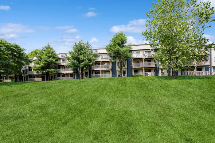 Charming apartment complex nestled among lush greenery, featuring spacious balconies and scenic views of well-manicured lawns under a bright blue sky.