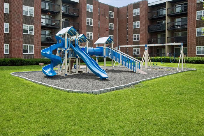 Charming apartment complex featuring a vibrant playground with blue slides and swings, surrounded by lush green lawns.