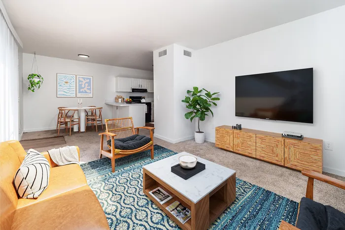 Modern living room with stylish decor, featuring a cozy mustard sofa, vibrant patterned rug, and a sleek wall-mounted TV beside a lush green plant. Open-concept design flows into dining area and kitchen with minimalist accents.