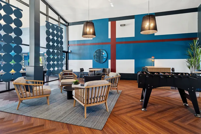 Modern communal lounge area with stylish wooden furniture, bold geometric wall art, and a sleek foosball table on elegant herringbone flooring.