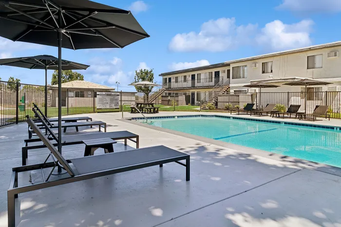 Modern apartment complex with a pristine swimming pool, sun loungers, and shaded seating areas, offering a luxurious outdoor retreat.