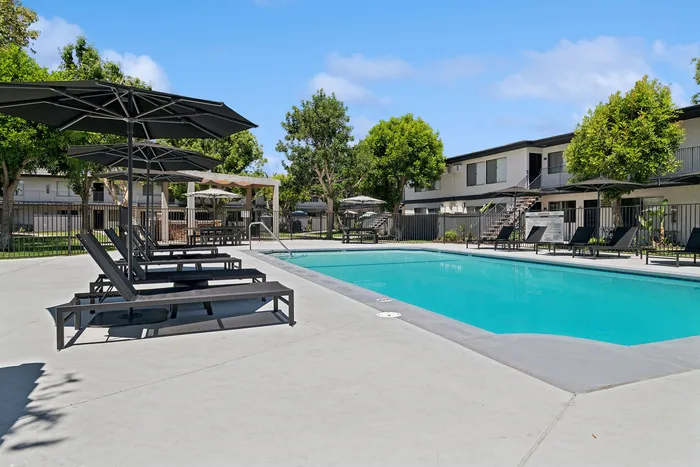 Inviting apartment complex pool area with stylish lounge chairs and umbrellas, surrounded by lush greenery and modern buildings.