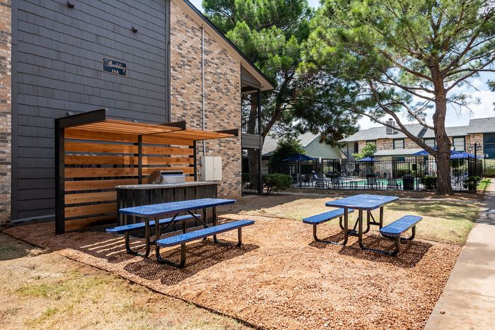 Charming outdoor area with blue picnic tables and a barbecue setup, shaded by trees next to a stylish apartment building and inviting pool area.