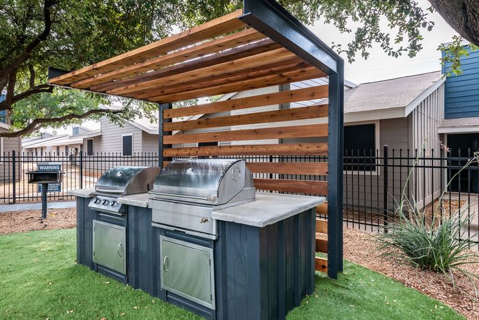 Chic outdoor grilling area with modern stainless steel BBQs under a stylish wooden pergola, surrounded by lush greenery.