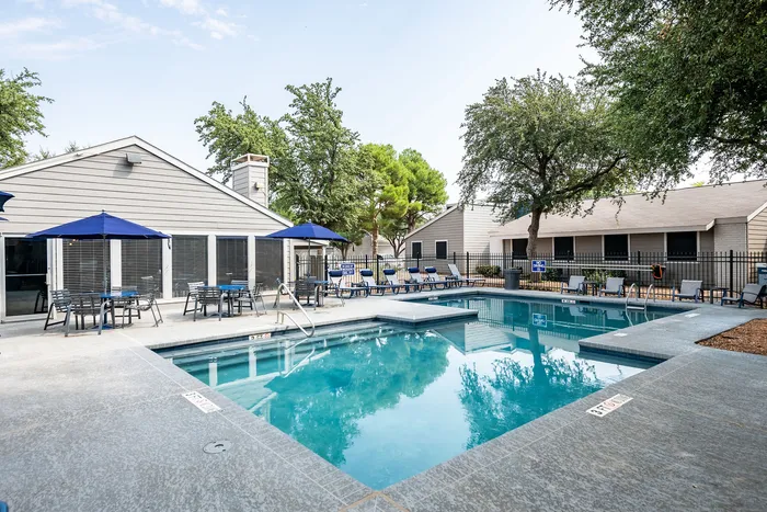 Inviting outdoor pool area with lounge chairs and blue umbrella-shaded tables surrounded by lush greenery, perfect for relaxation and social gatherings.