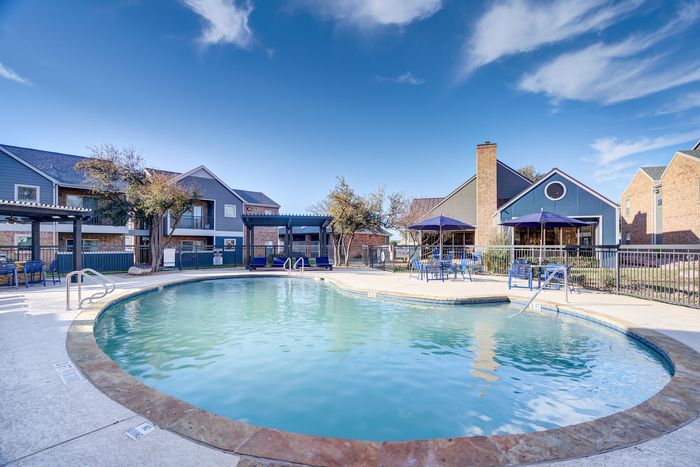 Inviting apartment complex with a sparkling pool, shaded seating, and charming architecture under a clear blue sky.