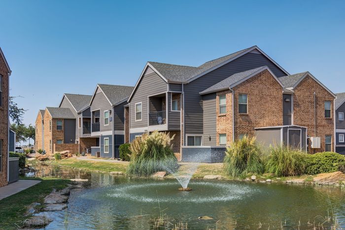 Charming apartment complex with modern architecture, lush landscaping, and a serene pond with a fountain.