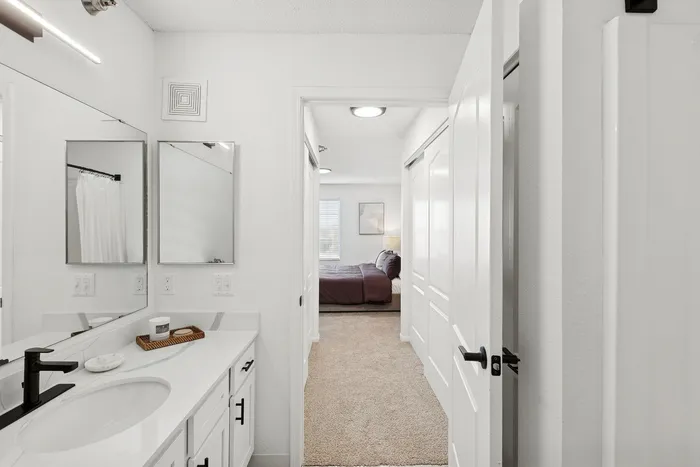 Elegant modern bathroom with sleek white vanity leading to a cozy bedroom, accentuated by plush carpeting and soft natural light.