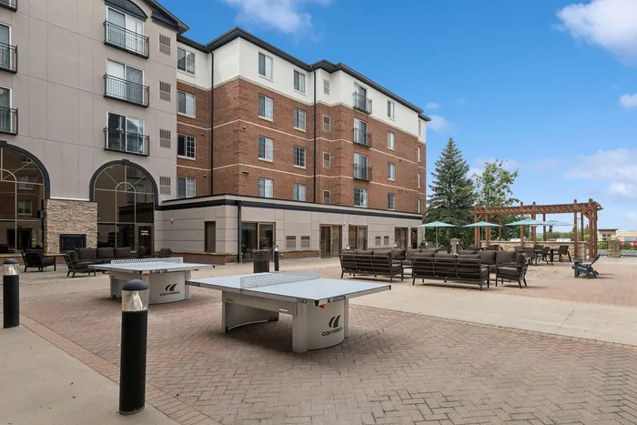 Vibrant apartment courtyard featuring ping pong tables, comfortable seating, and a cozy pergola area, all under a sunny blue sky.