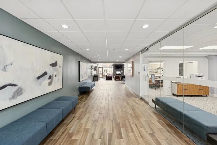Modern apartment lobby with sleek wood flooring, stylish gray sofas, abstract wall art, and glass office entrance for a welcoming atmosphere.