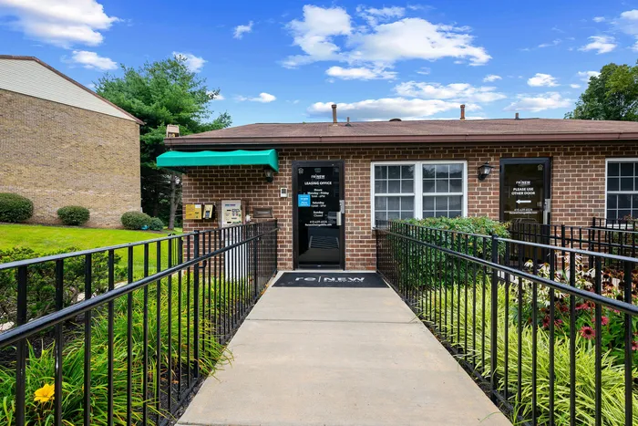 Charming leasing office entrance with vibrant greenery and a welcoming pathway, set against a bright, blue sky.
