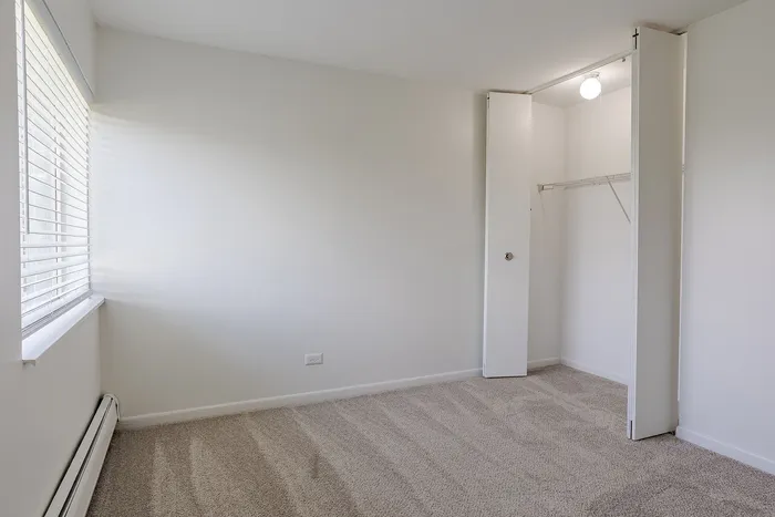 Bright and cozy bedroom with soft carpet flooring, open closet, and ample natural light from large window blinds.