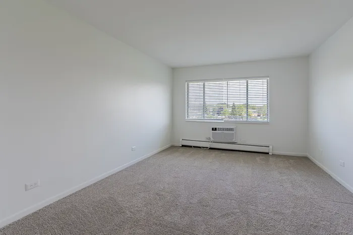 Bright and spacious empty room with plush carpet flooring, large window with blinds, and a built-in air conditioner.