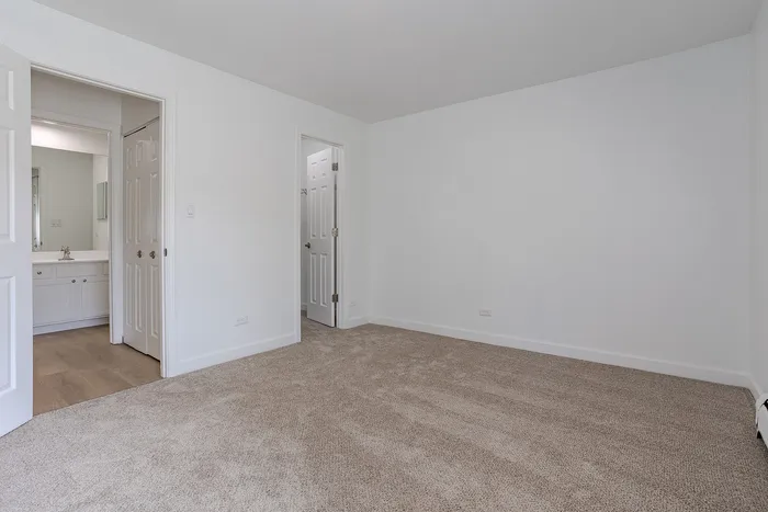 Pristine bedroom with plush carpeting, leading to a bright bathroom, showcasing modern design and ample natural light.