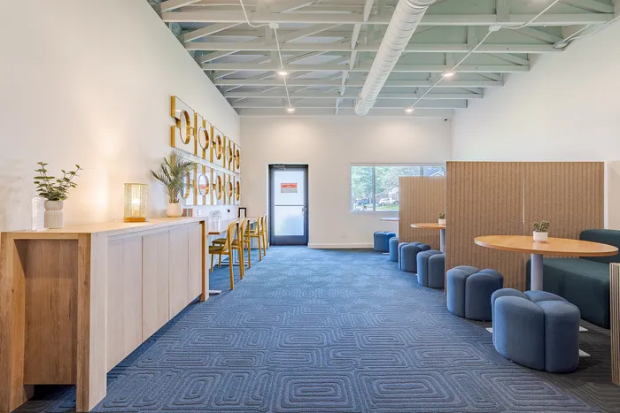 Modern communal space with stylish decor, featuring blue geometric carpet and cozy seating areas with vibrant natural lighting.
