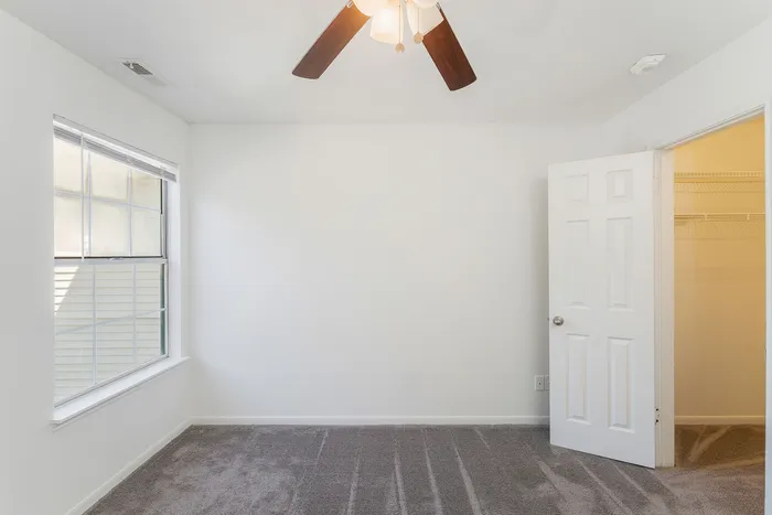 Bright, modern bedroom with plush carpet, large window, ceiling fan, and an inviting walk-in closet.
