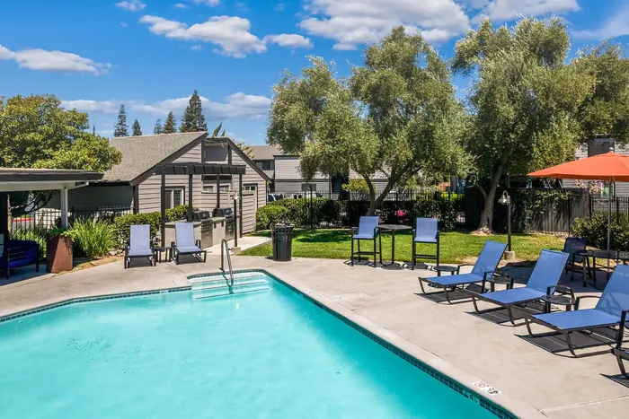 Inviting outdoor pool area with lounge chairs, shaded seating, and lush greenery under a bright blue sky.
