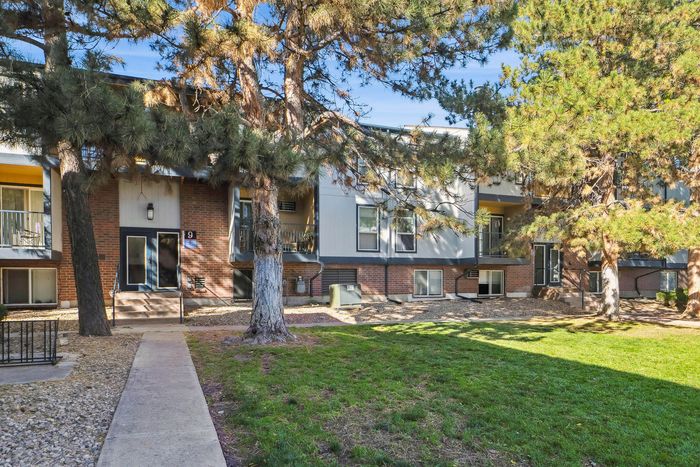 Charming apartment building with modern architecture surrounded by lush pine trees and a well-kept lawn, offering a serene living environment.