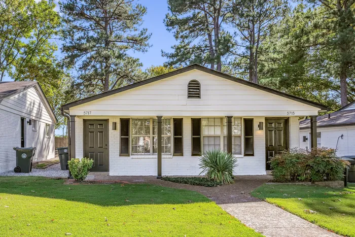 Charming white brick duplex with classic black doors and shutters, nestled among lush greenery and surrounded by mature trees, showcasing serene suburban living.