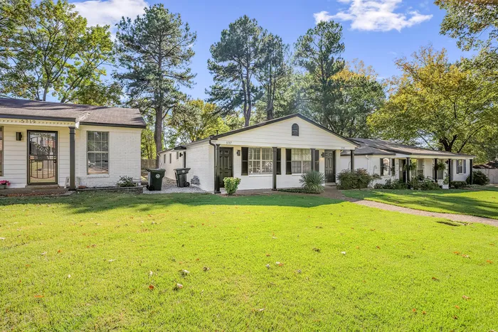 Charming suburban duplex with classic white brick facade, surrounded by lush green lawns and mature trees under a clear blue sky.