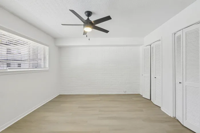 Bright and airy room featuring sleek light wood flooring, modern ceiling fan, and large window for natural light with white brick accent wall.