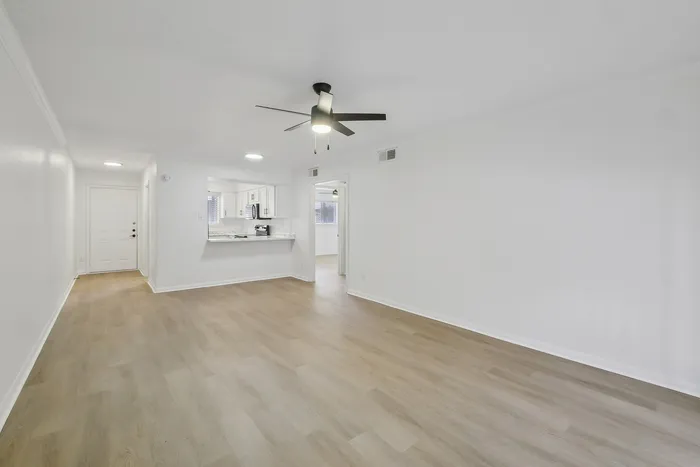 Bright open-concept living area with sleek wooden flooring, modern ceiling fan, and a view into a chic kitchen with white cabinetry.