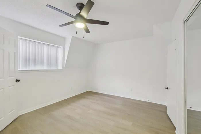 Bright modern bedroom with sleek wooden floors, large window with blinds, and a contemporary ceiling fan, perfect for a cozy retreat.