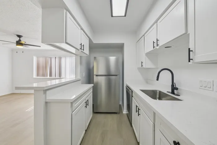 Sleek modern kitchen with white cabinetry, stainless steel fridge, and elegant countertop extending to a bright living area with ceiling fan.