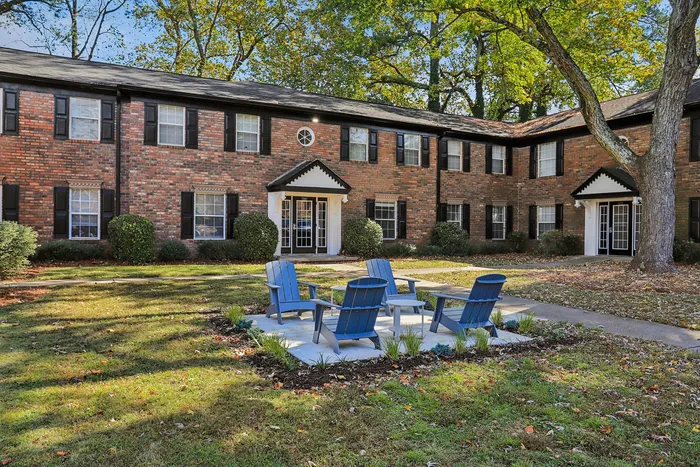 Charming brick apartment building with classic architecture, surrounded by vibrant greenery and featuring a cozy outdoor seating area with blue chairs for relaxation.