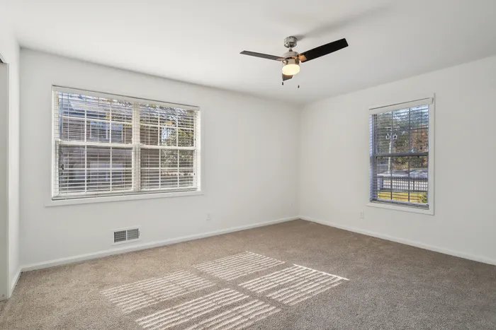 Bright and airy room featuring fresh white walls, plush carpeting, and large windows with a serene view, illuminated by a sleek ceiling fan light.