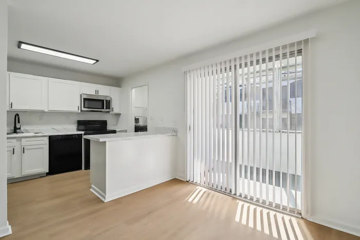 Bright kitchen with modern white cabinetry and sleek stainless steel appliances, seamlessly connected to a sunlit dining area with large window blinds.