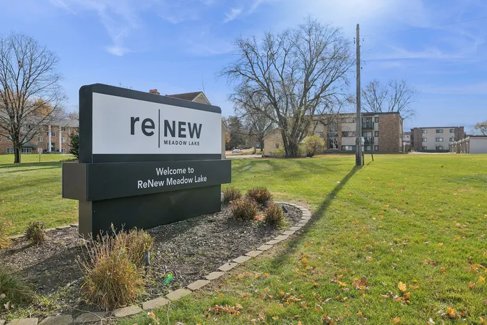 Welcome sign for ReNew Meadow Lake apartments surrounded by lush greenery and modern buildings under a clear blue sky.