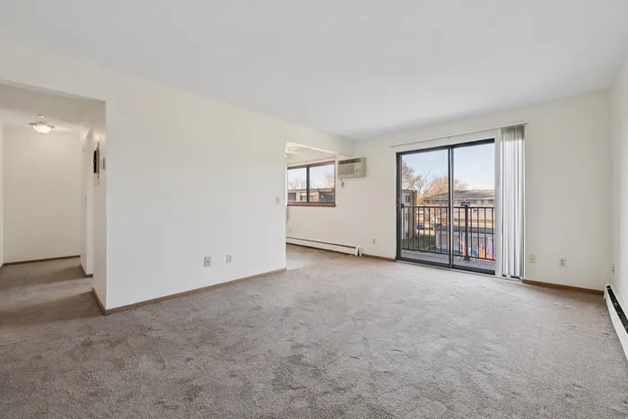 Bright and spacious living room with sliding glass doors leading to a balcony, featuring warm carpeted floors and natural light.