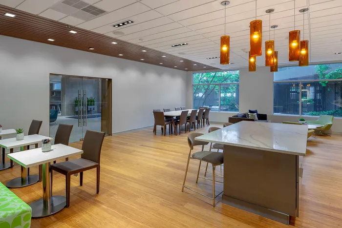Contemporary communal area featuring elegant dining tables, cozy seating, and modern pendant lighting with a view of lush greenery through large windows.