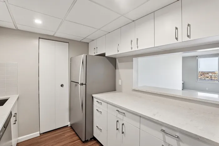 Modern kitchen with sleek white cabinetry, a stainless steel refrigerator, and a pass-through to a light-filled room with city views.