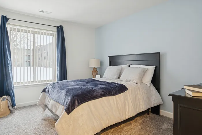 Bright and cozy bedroom with a large window, elegant blue curtains, and a plush bed with soft pillows and a stylish dark headboard.